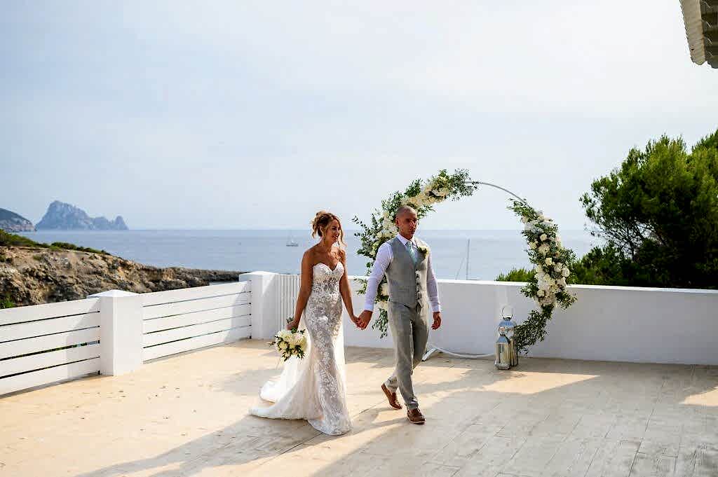 A bride and groom holding hands and walking on a terrace overlooking the sea, with a floral arch in the background. The groom is in a light grey suit, and the bride is in a strapless white lace wedding dress.